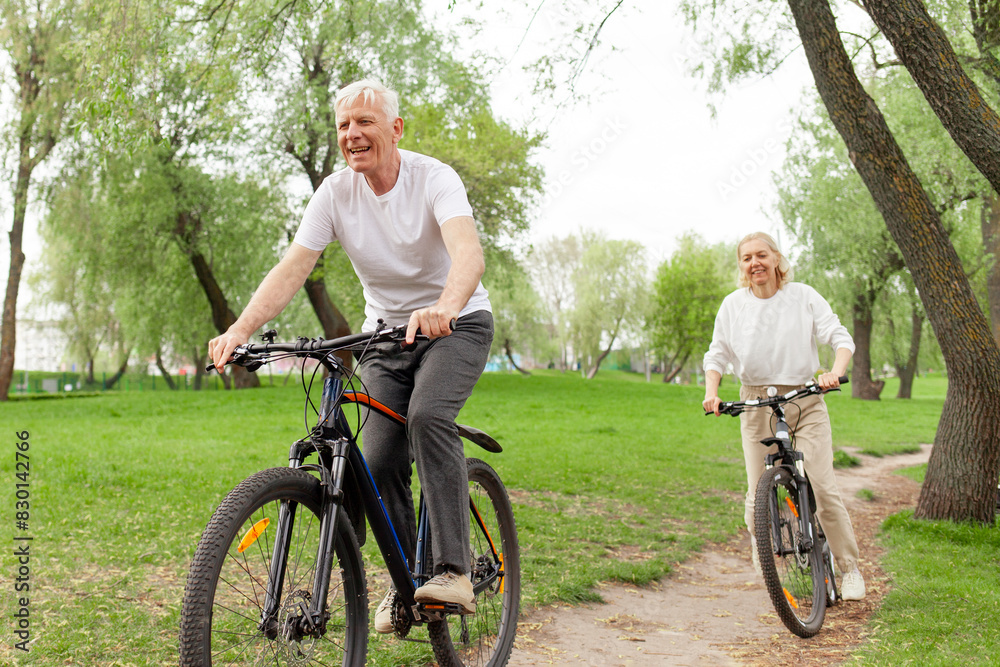 Obraz premium elderly senior couple rides bicycle in the park in the summer and smiles, old gray-haired man and woman are actively resting outdoors, old people practice cycling in forest