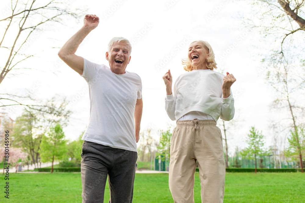 elderly senior couple man and woman rejoice in victory and jump in the park outdoors, gray-haired grandparents celebrate luck and success and rejoice in nature