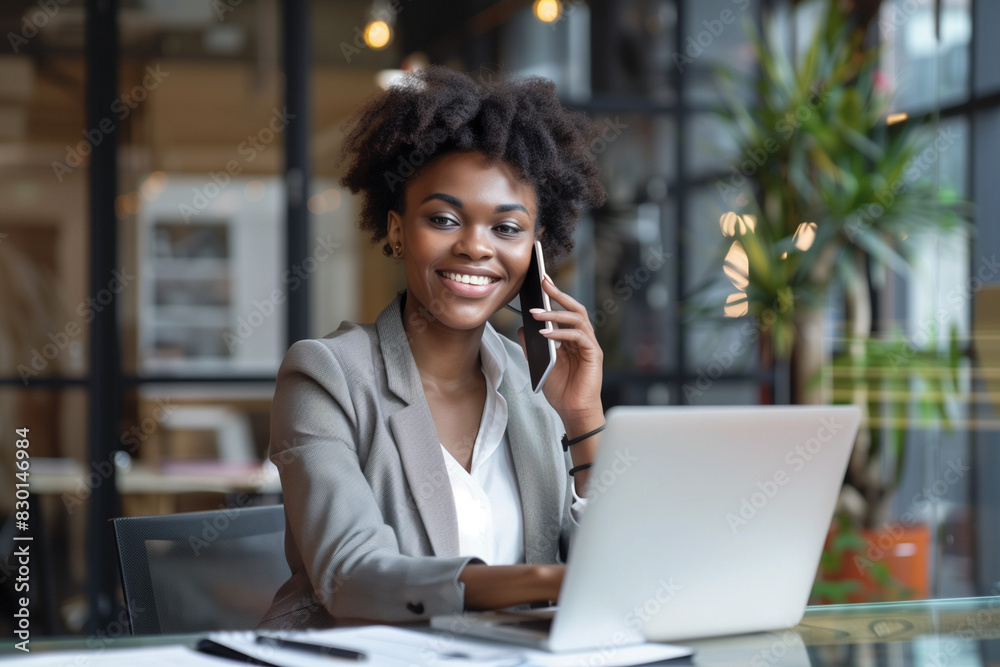 Smiling Businesswoman on the Phone. Cheerful African American businesswoman wearing glasses, talking on the phone while working on her laptop in a modern office environment. Smiling Businesswoman on the Phone. Cheerful African American businesswoman wearing glasses, talking on the phone while working on her laptop in a modern office environment.