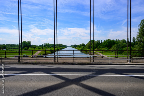 Foto van een europese brug met de sphalt, brugstructuur in de voorgrond en het kanaal en bossen op de achtergrond. Blauwe lucht witte wolken.