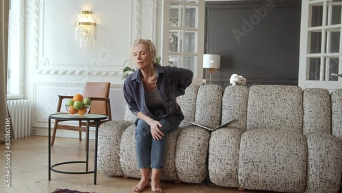 Woman in 60s sitting on couch in living room with laptop. She works or enjoys leisure online activities. tries to stand up and feels unexpected sharp back pain causes her to frown and hold her back