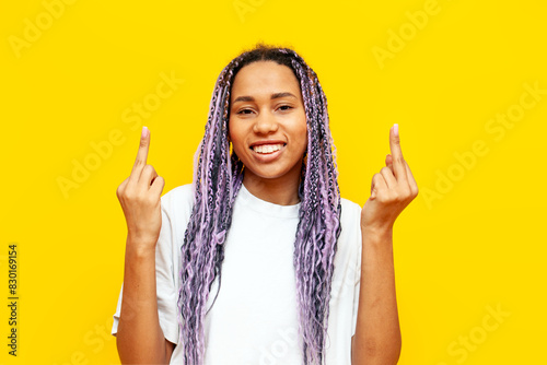 cheerful african american woman with colored dreadlocks showing middle finger on yellow isolated background, hipster girl with purple braids and unique hairstyle showing fuck gesture photo