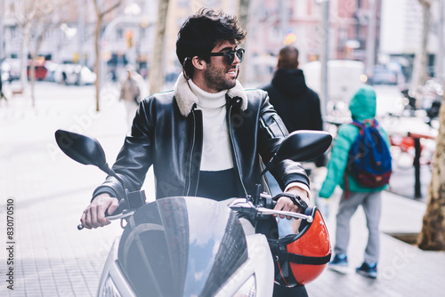 Cheerful hipster guy in black sunglasses parking own motorcycle on pavement in urban setting.Bearded biker in spectacles and leater jacket sitting on motorbike while looking away photo