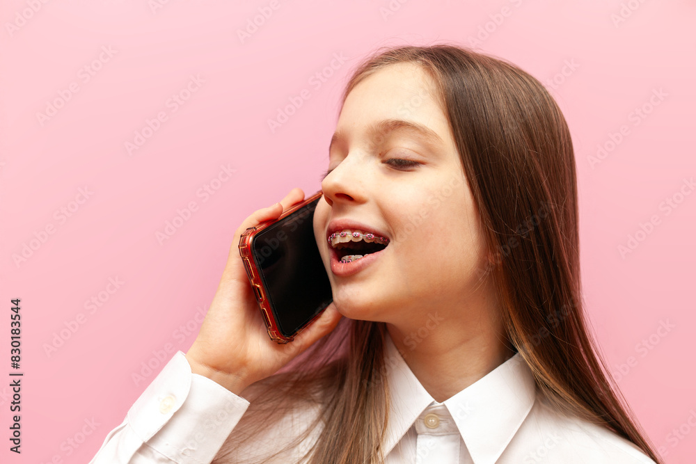 cheerful little teenage girl with braces talking on the phone on a pink isolated background, child communicating via mobile phone cheerful little teenage girl with braces talking on the phone on a pink isolated background, child communicating via mobile phone