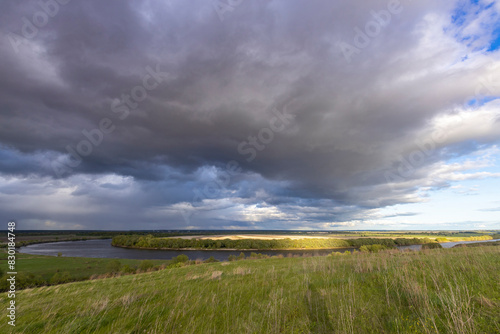 A serene meadow overlooks a winding river under a dramatic sky filled with storm clouds and patches of blue, capturing the beauty of nature's ever-changing weather patterns