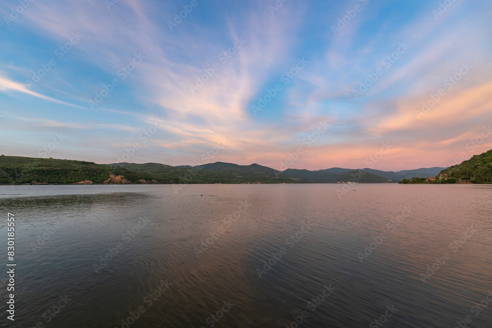 A serene sunset over a tranquil lake with rippled water, mountains in the distance, and a clear sky with soft clouds