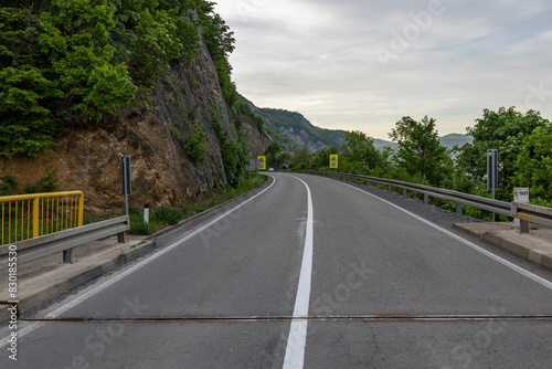captures a serene  overcast day on a mountain road. The asphalt road curves gently to the right  bordered by a yellow and white guardrail on the left and a series of safety barriers on the right.