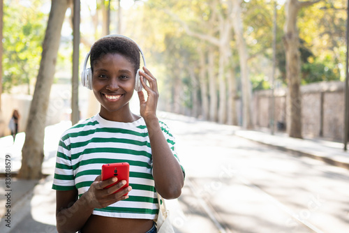 Young African woman listening to music with headphones and wireless red cell pone at city park  photo