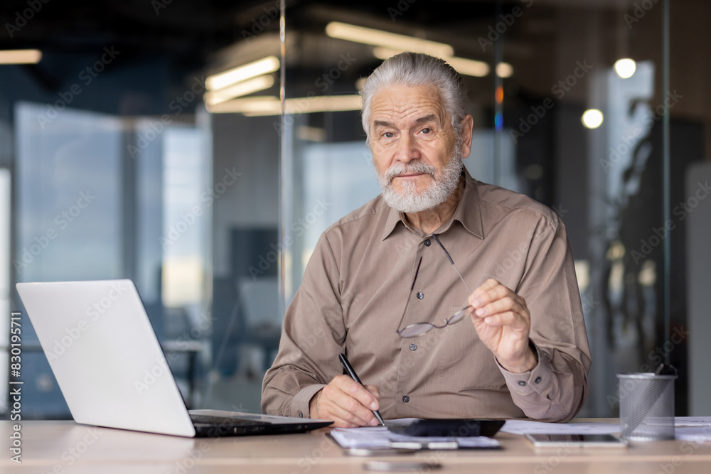 The senior professional is seated at his office desk with a laptop, holding his glasses in a modern workplace setting The senior professional is seated at his office desk with a laptop, holding his glasses in a modern workplace setting