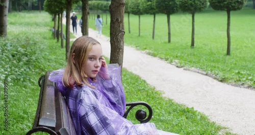 Teen girl wearing raincoat sitting on bench in summer landscaped park at rainy warm weather. Blonde teenager rubbing eyes boring on street on nature. Female waiting somebody looking by sides.