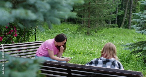 Mom daughter browsing smartphones sitting on benches in forest park in summer. Pastime leisure, family digital entertainment together. Woman girl surfing online in phones. Internet addiction. 