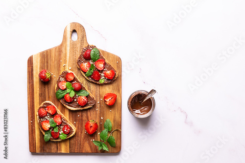 Sweet homemade toasts with chocolate paste, nuts, srtawberry and aromatic mint leaves on the serving board on white marble background. Tasty summer dessert. photo