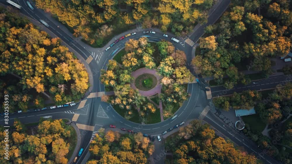 Top down time-lapse aerial view of a park with a roundabout intersection with slow camera rotation, beautiful sunset illuminates trees. Cars move fast along the road during an autumn traffic jam.