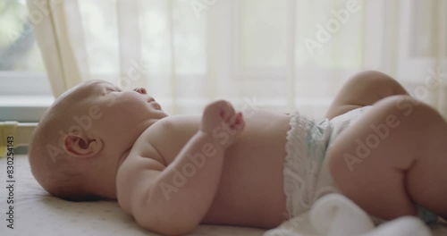 Cute infant baby in diaper lying in bed and looking at camera. Close up of sweet beautiful newborn baby lying on bed sheets, plays touches toes laughs smile looks at camera. photo