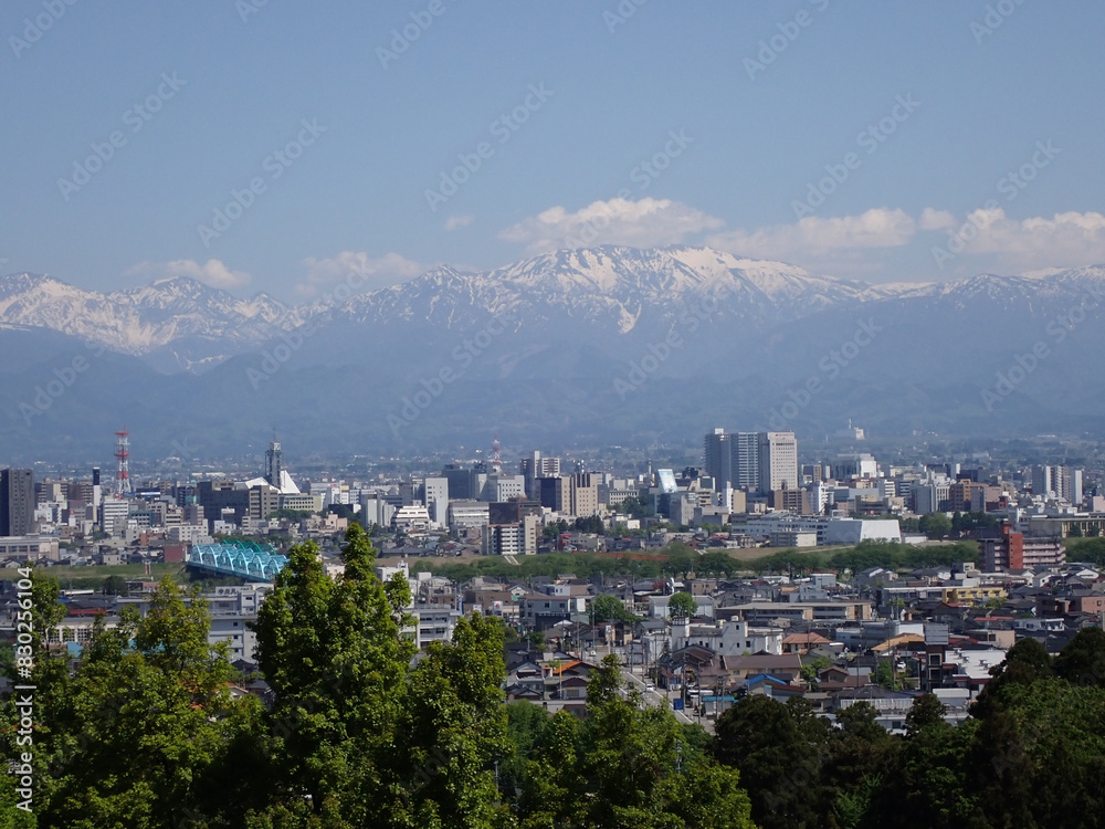 Obraz premium 5月新緑の木々と富山市街地の後方に見える残雪の北アルプス薬師岳