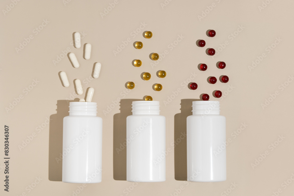 Fototapeta premium Three white mockup jars with natural white, yellow and red pills capsules on beige isolated background. The concept of pharmacy, health, medicines and dietary supplements. Image for your design.