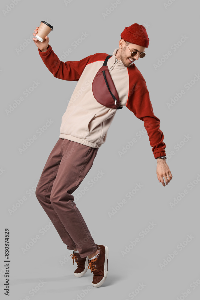 Handsome smiling young man in stylish clothes with sunglasses, bag and cup of coffee posing on grey background Handsome smiling young man in stylish clothes with sunglasses, bag and cup of coffee posing on grey background