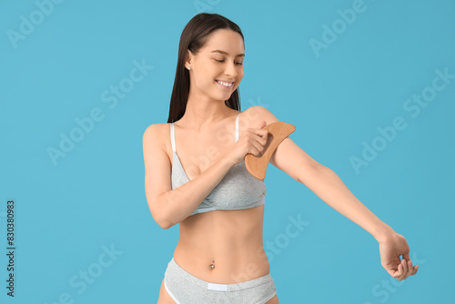 Young woman in underwear massaging her arm with wooden scraper on blue background © Pixel-Shot
