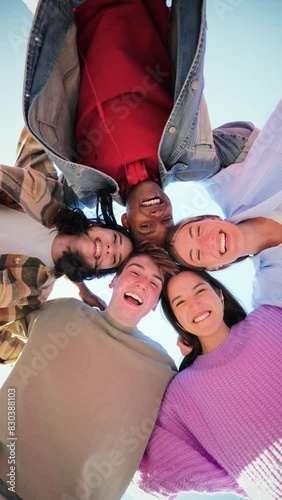 Vertical low angle view of a group of multiracial young friends hugging in a circle, smiling and looking at camera with a positive expression. Team of teenagers laughing and ambrecing each others