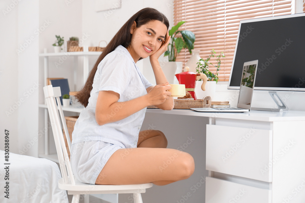 Pretty young woman with cup of hot coffee and laptop sitting at table in bedroom