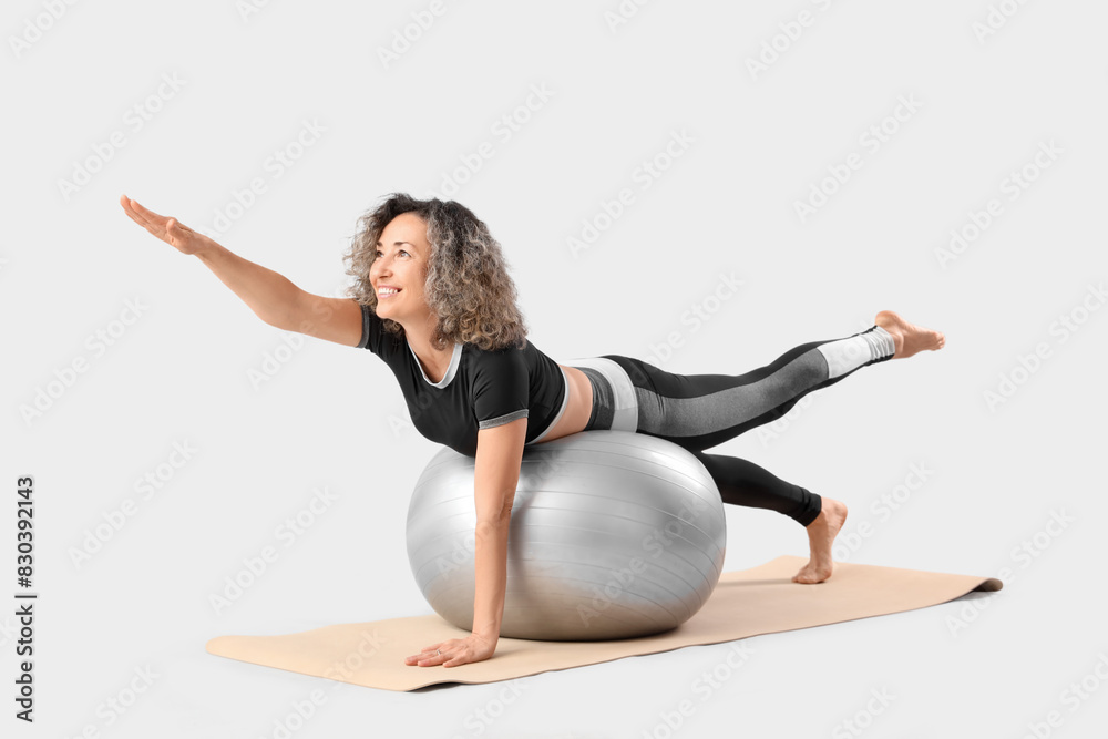 Sporty mature woman doing yoga on fitball against light background