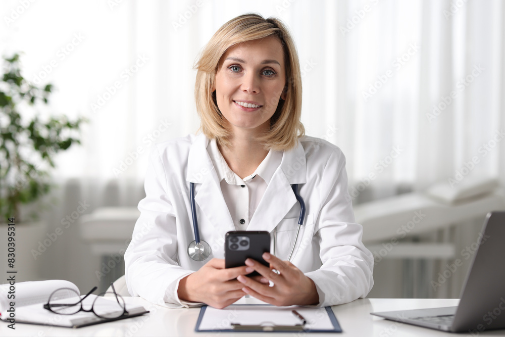 Smiling doctor with smartphone having online consultation at table in office Smiling doctor with smartphone having online consultation at table in office