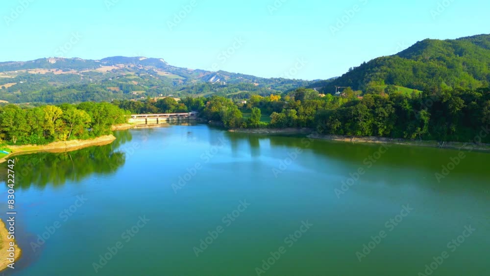 Smooth aerial push-in shot from Lago di San Ruffino, calm lake waters skimming vegetation-filled shores and a bridge in the distance, with a vision of typical Marche hillscapes, their trees and fields Smooth aerial push-in shot from Lago di San Ruffino, calm lake waters skimming vegetation-filled shores and a bridge in the distance, with a vision of typical Marche hillscapes, their trees and fields