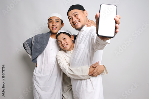 Happy Asian Muslim Man With His Two Sons Showing Blank Screen Smartphone For Mockup Or Text photo