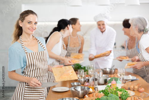 Young participiant of cooking master class holding cutting board with raw chicken breast in her hands standing around other female members