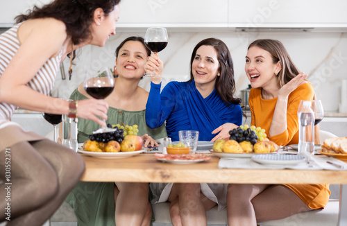 Relaxed adult female friends having fun at intimate house party, sharing stories and laughing gathered around table with wine and appetizers in cozy kitchen