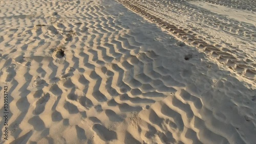 an old scruffy soccer ball rolling along beach sand with tire imprints in the sand kicked by a male with a black shoe
