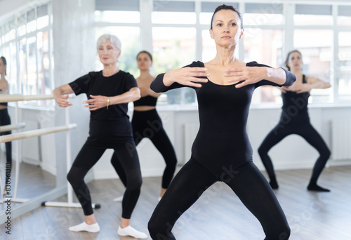 Motivated fit woman, participating in ballet class, practicing basic movements with female group of various ages in bright choreography studio with natural lighting..