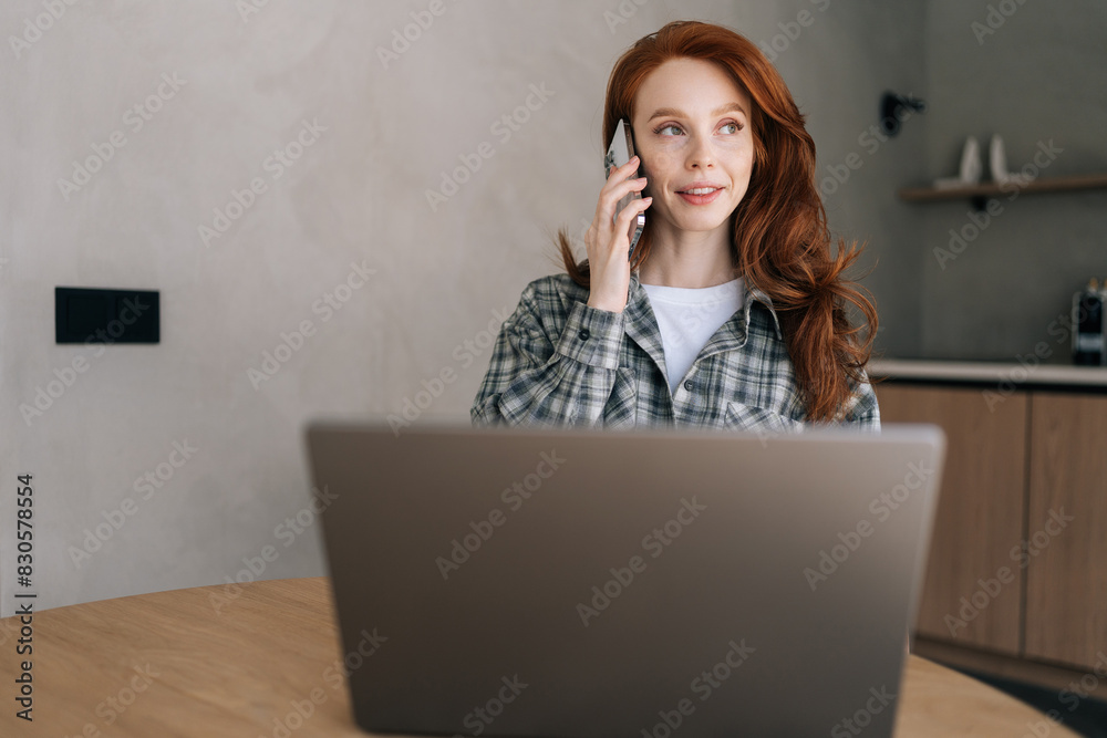 Portrait of attractive female manager sitting at home desk in front of laptop holding mobile phone, make pleasant business or informal call. Happy web designer consulting client about online project. Portrait of attractive female manager sitting at home desk in front of laptop holding mobile phone, make pleasant business or informal call. Happy web designer consulting client about online project.