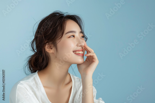 a beautiful Asian woman with medium hair in a white blouse touching her nose and smiling while showing off the side of her face against a light blue background