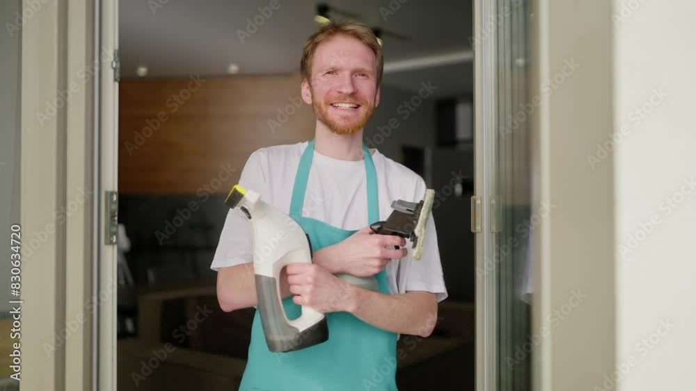 Portrait of a confident blond man with a beard in a white T-shirt and with a blue apron a cleaner holds in his hands two vacuum cleaners for windows during his cleaning in a modern apartment on call Portrait of a confident blond man with a beard in a white T-shirt and with a blue apron a cleaner holds in his hands two vacuum cleaners for windows during his cleaning in a modern apartment on call