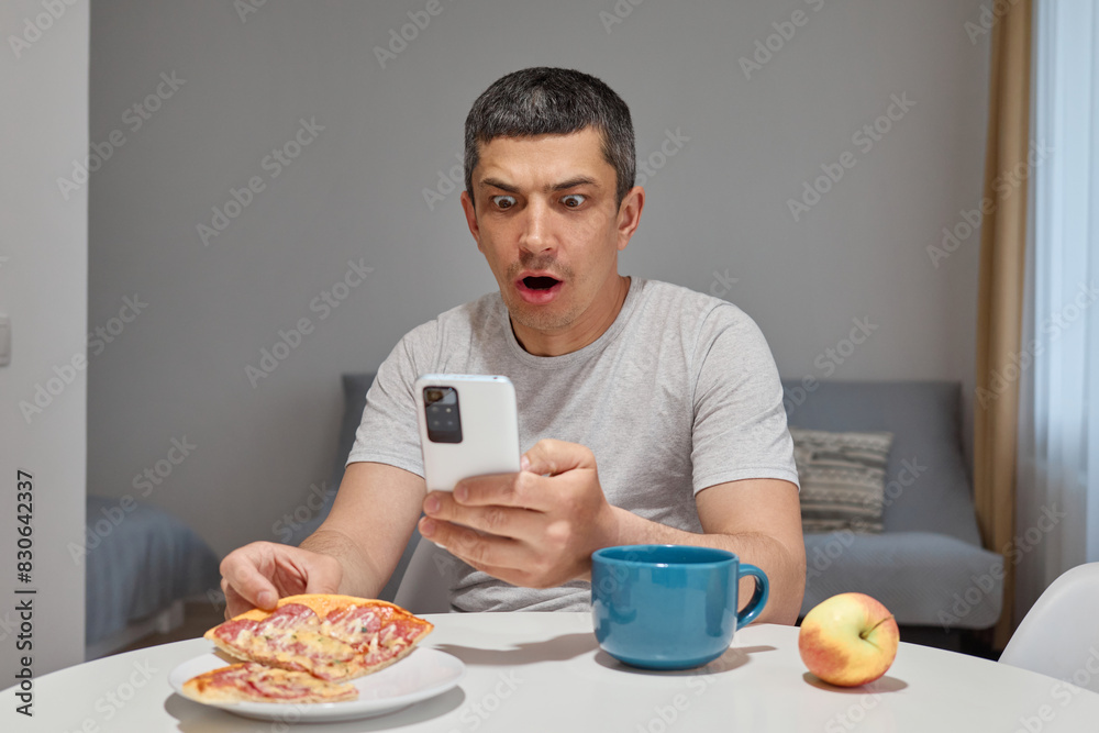 Shocked surprised astonished Caucasian man wearing T-shirt sitting at table in home interior holding smartphone reading internet news looking at device screen with big eyes Shocked surprised astonished Caucasian man wearing T-shirt sitting at table in home interior holding smartphone reading internet news looking at device screen with big eyes