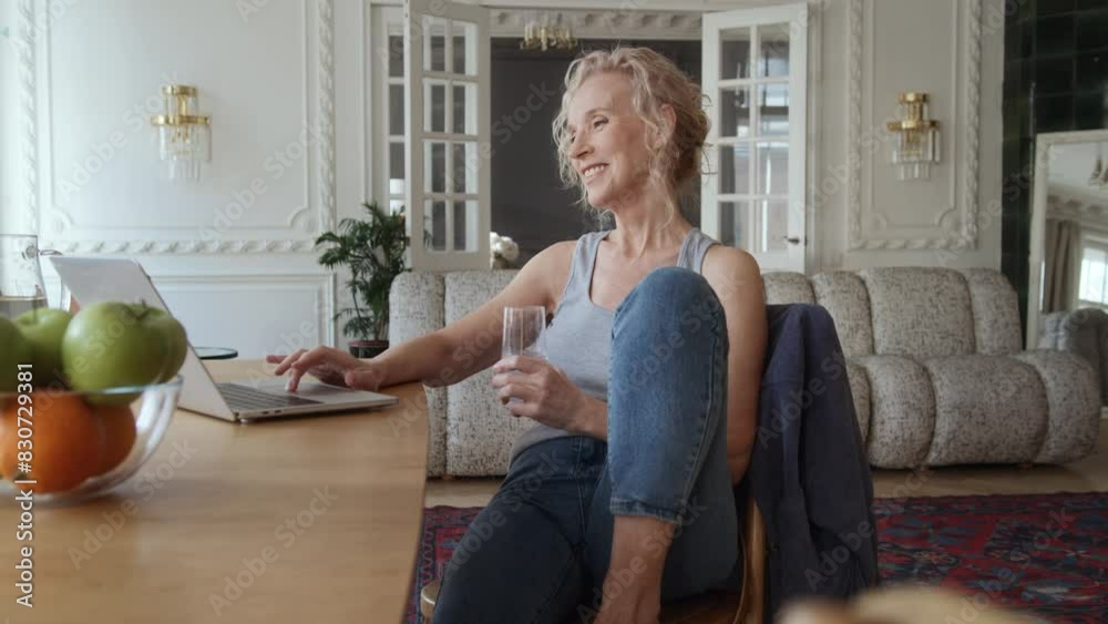 Senior woman sitting at table in living room with laptop. She works remotely or enjoys leisure activities online shopping typing on keyboard. Satisfaction and fulfillment in the older woman life. Senior woman sitting at table in living room with laptop. She works remotely or enjoys leisure activities online shopping typing on keyboard. Satisfaction and fulfillment in the older woman life.