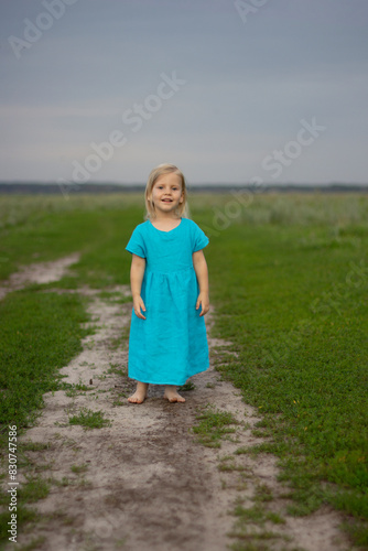 Little happy beautiful blonde girl in a blue linen sundress against the background of a green meadow, live emotions, smile. sunny day