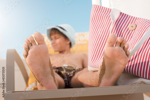 Kid feet on sunbeds on sand beach, boy in hat is smiling vacation concept