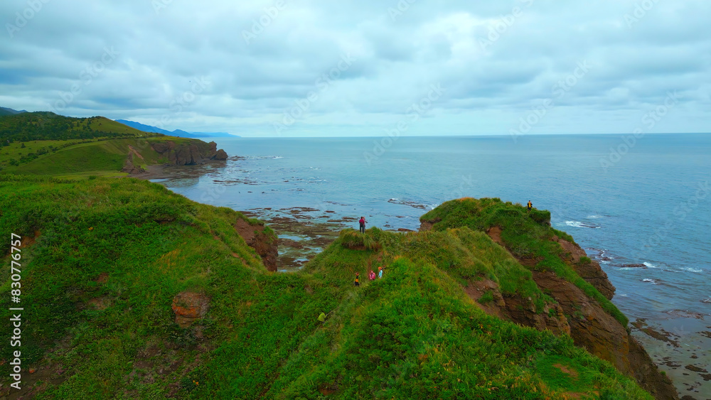 Top view of tourists on rocky coast trail with green grass. Clip. Beautiful landscape of rocky coast with green grass and tourists on hike. Tourists ride on edge of rocky coast overlooking sea on