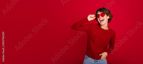 Banner image of a gorgeous smiling excited trendy wear woman in glasses in heart shape isolated on a red bright wall. Girl in good mood and with emotions of love © My Ocean studio