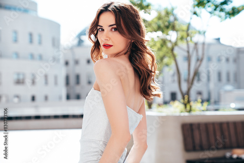 Young beautiful brunette female in trendy evening silver dress. Sexy carefree woman posing in the street at sunny day. Fashionable model with evening bright makeup and red lips  outdoors