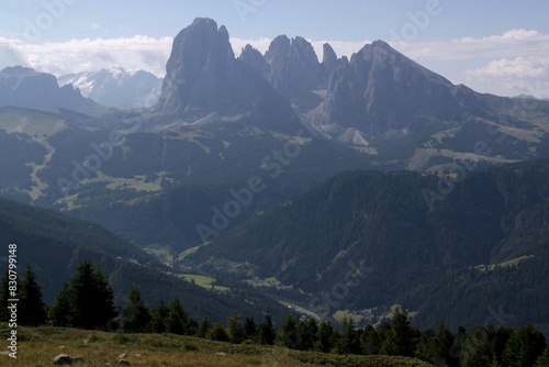 Panoramic view of the landscape with the Sassolungo mountain massif in the morning fog and illuminated by the morning sun with a meadow and pine trees in the Dolomites, South Tyrol region, Italy