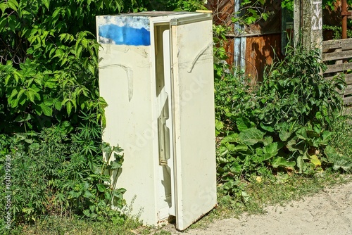 one old dirty closed discarded rusty white electric closed iron refrigerator stands on the ground near green bushes  during the day on the street photo