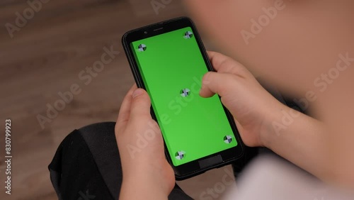 Close-up of a little boy holding a phone with a green screen sitting at home on the couch. 