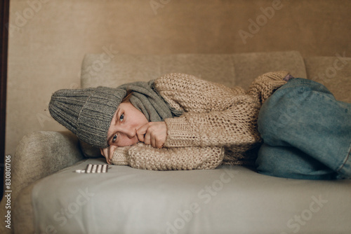 Young woman wearing scarf and wool hat has flu ill sick disease cold at home indoor on sofa