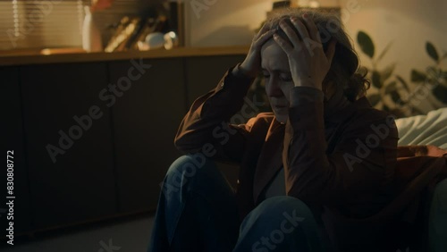 Side footage of tired senior woman sitting on floor in dark room alone and covering face with hands in despair after receiving bad news at night
