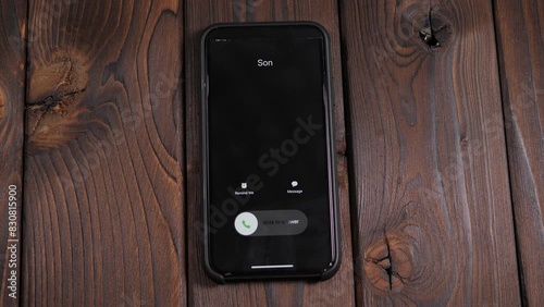 A close-up of an incoming call from his son on a smartphone screen on a wooden background, a man answers the call.