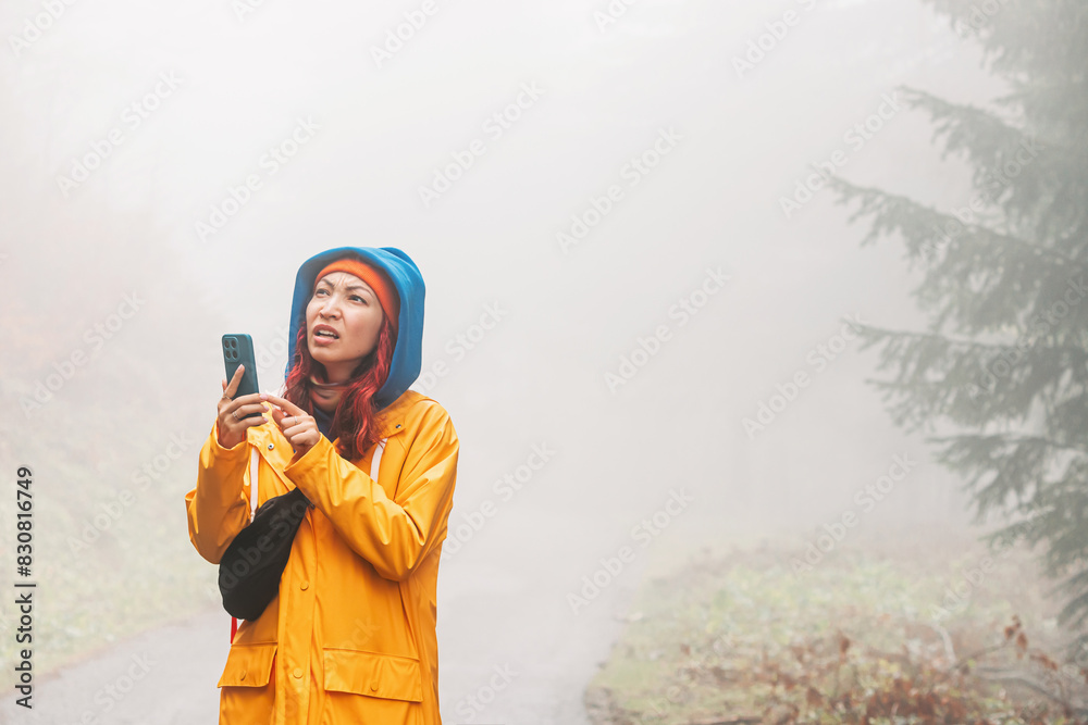 © EdNurg - Lost in the dense fog of the mountains, a worried woman hiker uses her smartphone for navigation, hoping for a strong battery and reliable connection to guide her back.