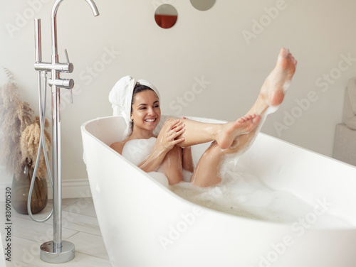 Positive Caucasian woman enjoying hot bath lying in tub at home and laughing in good mood. Pretty young female bathing in bathtub with soap foam. Lady with long legs taking a bath photo